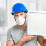 Man wearing a blue hard hat and face mask holding an air conditioner filter, emphasizing the importance of clean filters for improved indoor air quality and AC maintenance in Vancouver homes.